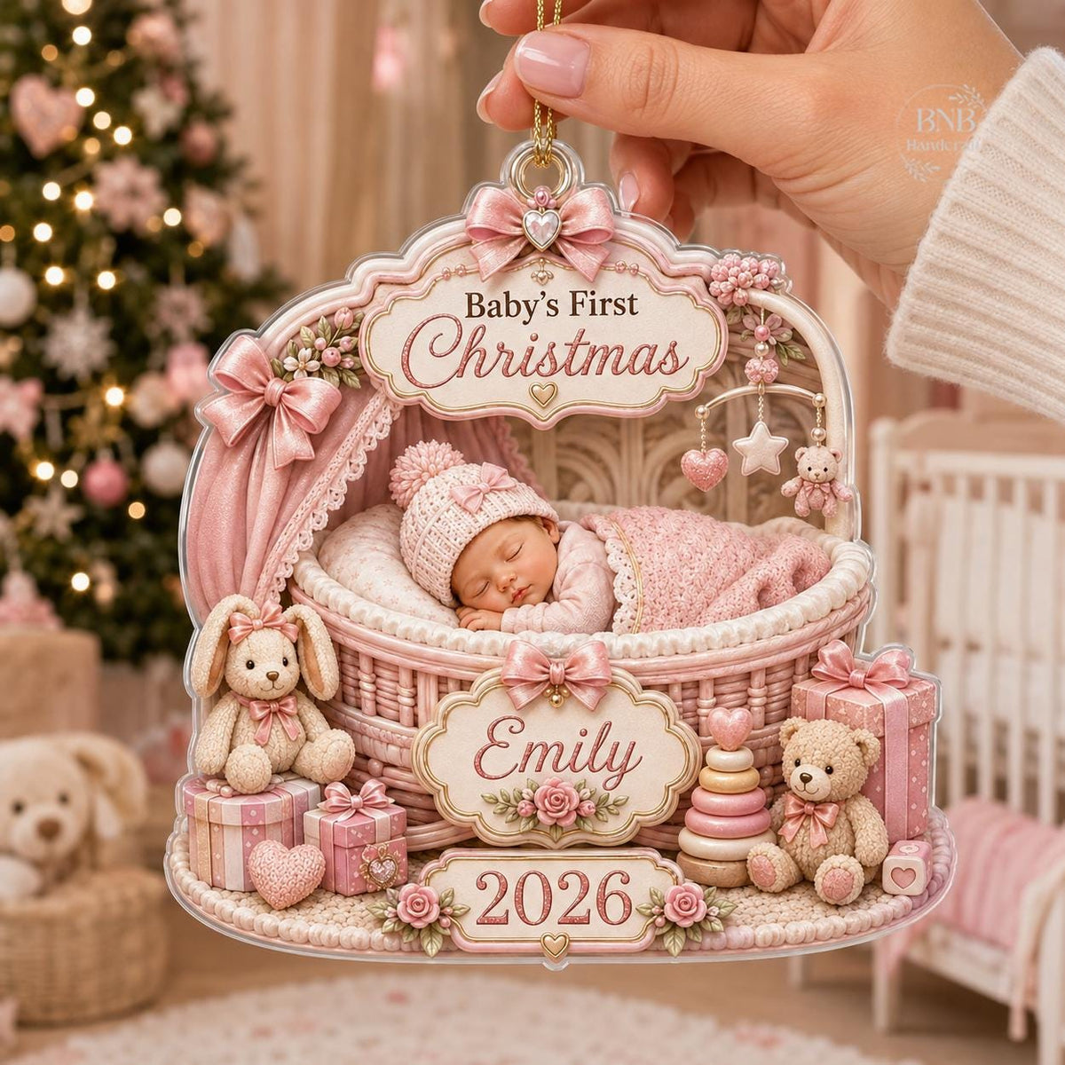 May include: A decorative acrylic ornament featuring a sleeping baby in a pink wicker bassinet. The text reads Baby's First Christmas, Emily, and 2026. It is adorned with pink bows, a plush bunny, a teddy bear, a stacking toy, and gift boxes.