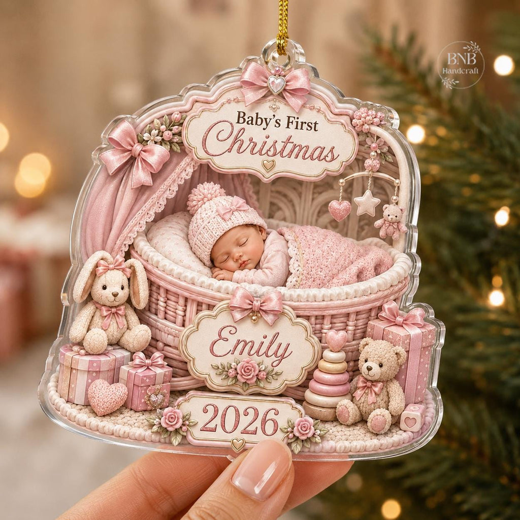May include: A clear acrylic Christmas ornament featuring a baby sleeping in a pink wicker bassinet. The text reads Baby's First Christmas, Emily, and 2026. The design includes a pink bunny, a teddy bear, gift boxes, and floral accents.