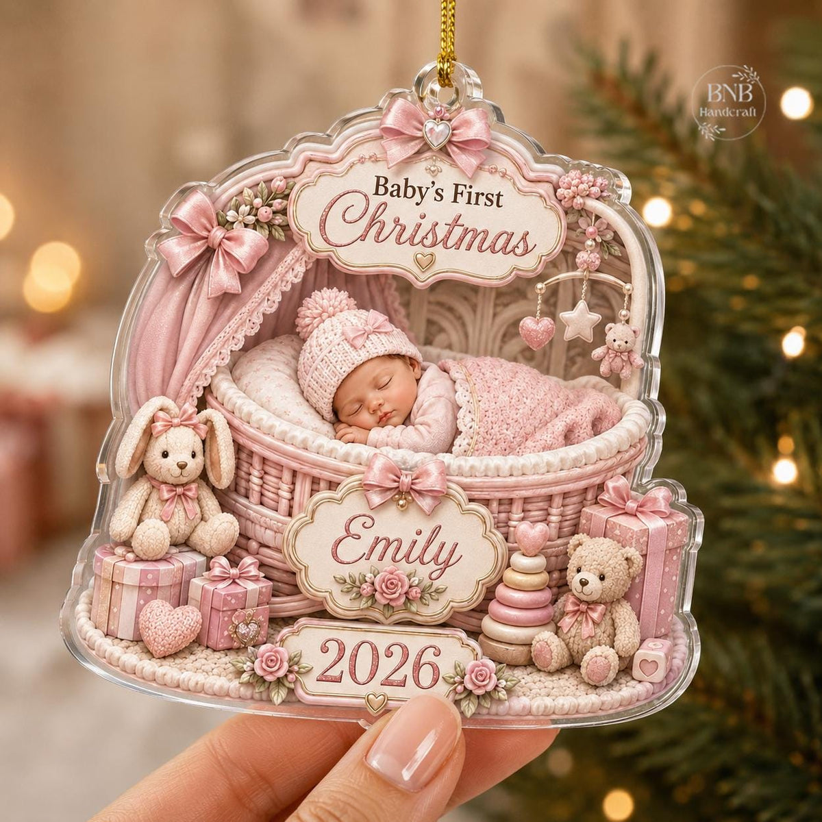 May include: A clear acrylic Christmas ornament featuring a baby sleeping in a pink wicker bassinet. The text reads Baby's First Christmas, Emily, and 2026. The design includes a pink bunny, a teddy bear, gift boxes, and floral accents.