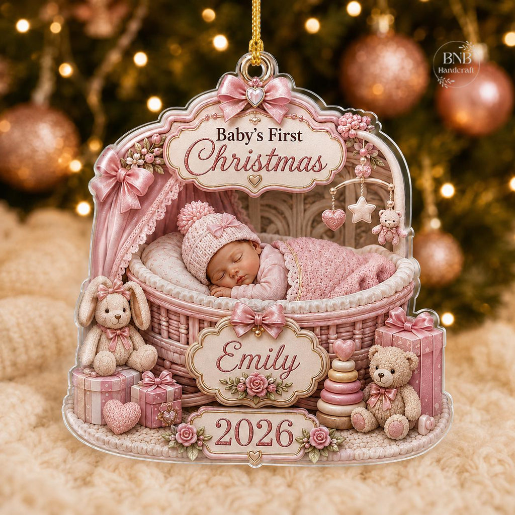 May include: A decorative acrylic ornament featuring a sleeping baby in a pink wicker bassinet. The text reads Baby's First Christmas, Emily, and 2026. The design includes pink bows, a plush bunny, a teddy bear, gift boxes, and a stacking toy.