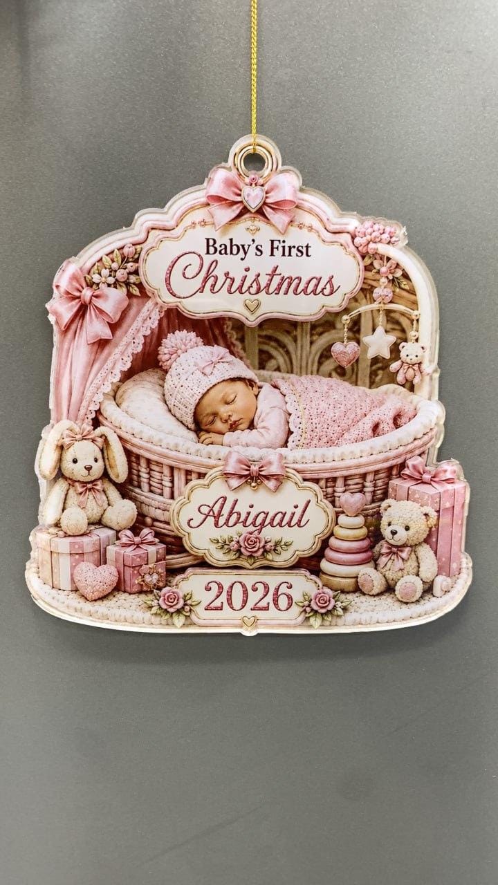 May include: A decorative hanging ornament featuring a sleeping infant in a wicker bassinet, pink bows, teddy bears, and wrapped gifts. The text reads Baby's First Christmas, Abigail, and 2026. The design is rendered in soft pink and cream tones.