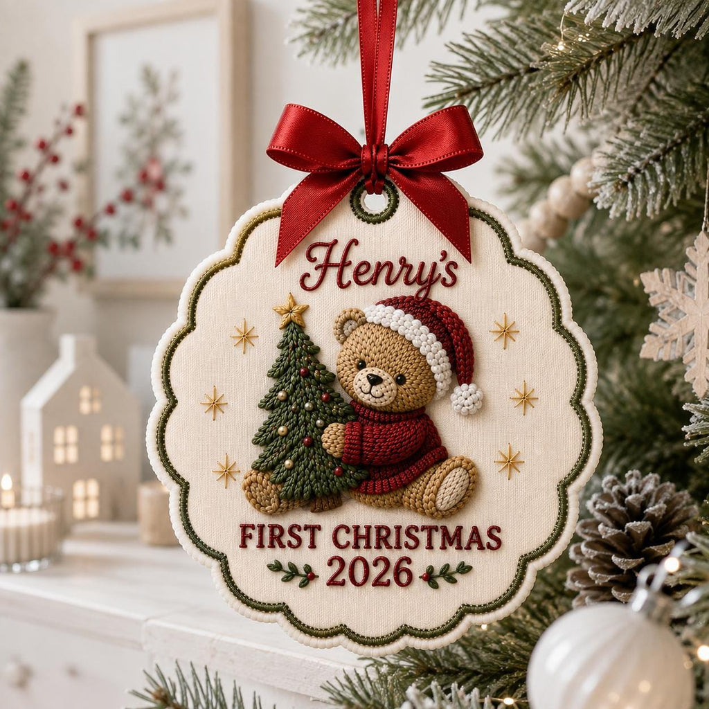 May include: A scalloped, cream-colored fabric ornament featuring an embroidered brown teddy bear wearing a red sweater and Santa hat, holding a green Christmas tree. The text reads Henry's FIRST CHRISTMAS 2026. It is topped with a red satin ribbon bow.