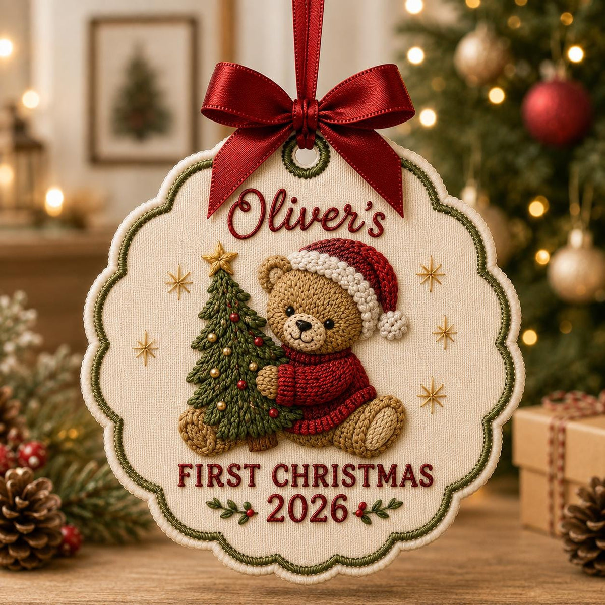 May include: A scalloped, cream-colored fabric Christmas ornament featuring an embroidered brown teddy bear in a red sweater and Santa hat holding a green Christmas tree. The text reads Oliver's FIRST CHRISTMAS 2026. A red satin ribbon bow is at the top.