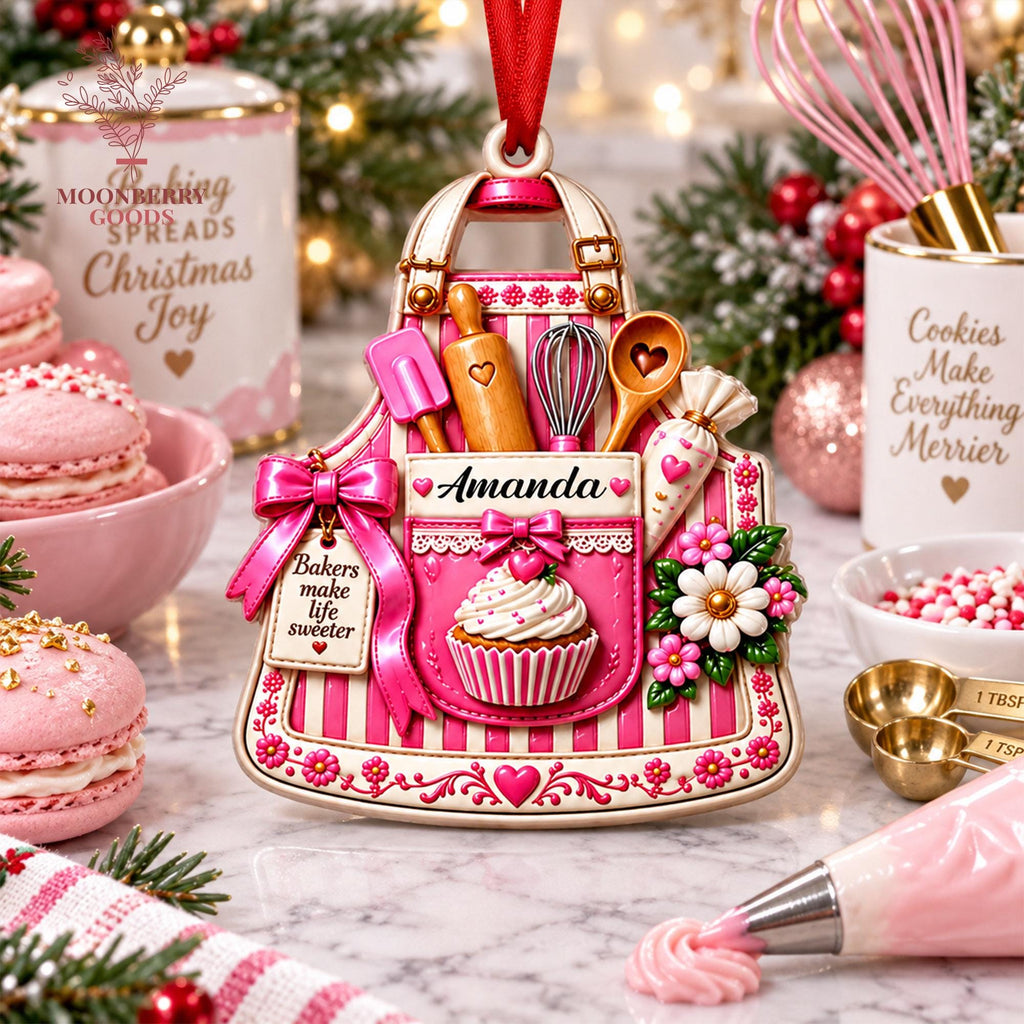 May include: A decorative ornament shaped like a pink and white striped apron featuring a cupcake, a rolling pin, a whisk, a wooden spoon, and a piping bag. The apron displays the name Amanda and a tag reading Bakers make life sweeter.