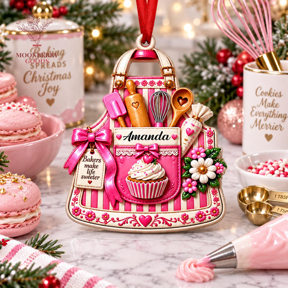 May include: A decorative ornament shaped like a pink and white striped apron featuring a cupcake, a rolling pin, a whisk, a wooden spoon, and a piping bag. The apron displays the name Amanda and a tag reading Bakers make life sweeter.