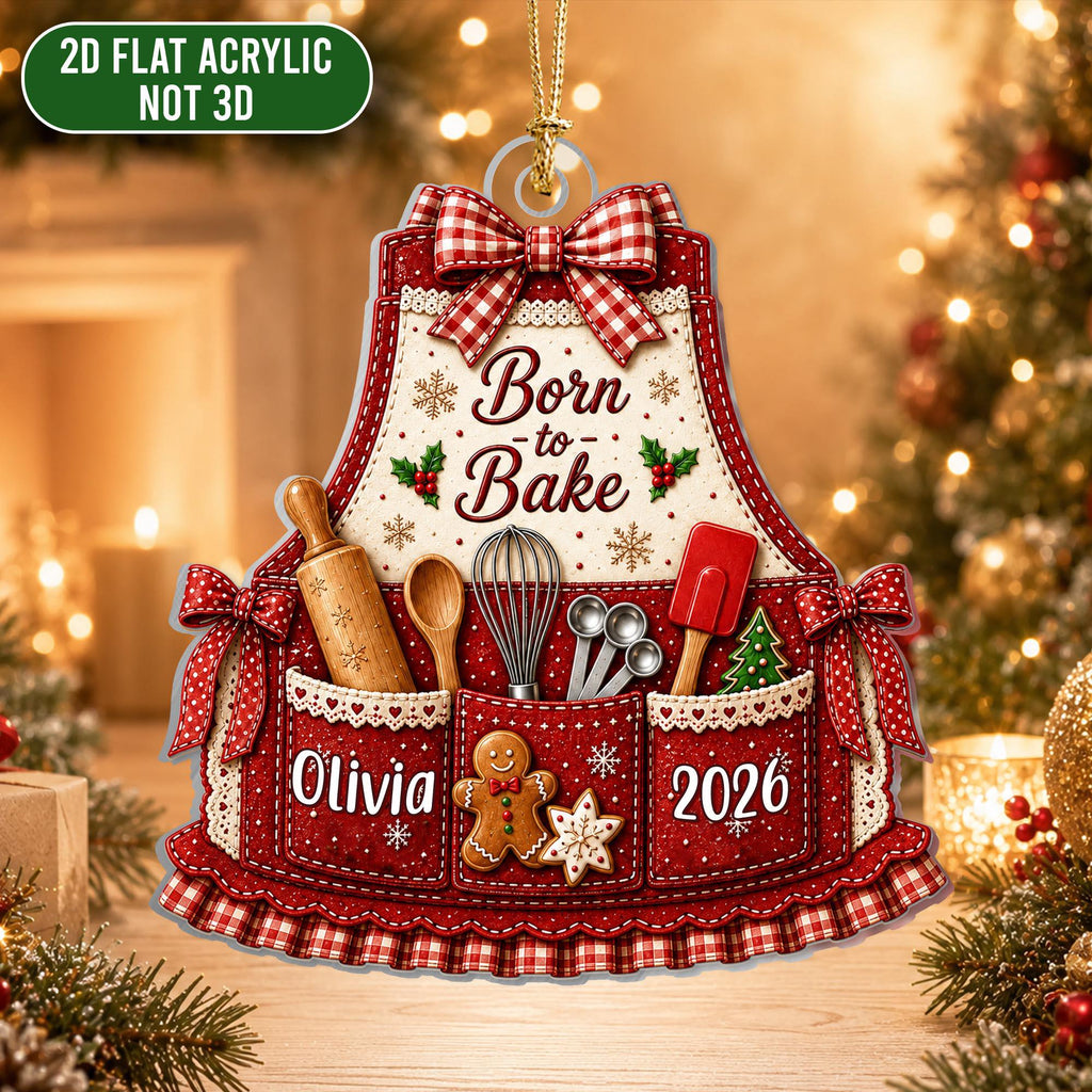 May include: A 2D flat acrylic ornament shaped like an apron with a red and white gingham bow, featuring the text Born to Bake, a rolling pin, whisk, measuring spoons, spatula, a gingerbread man, the name Olivia, and the year 2026.