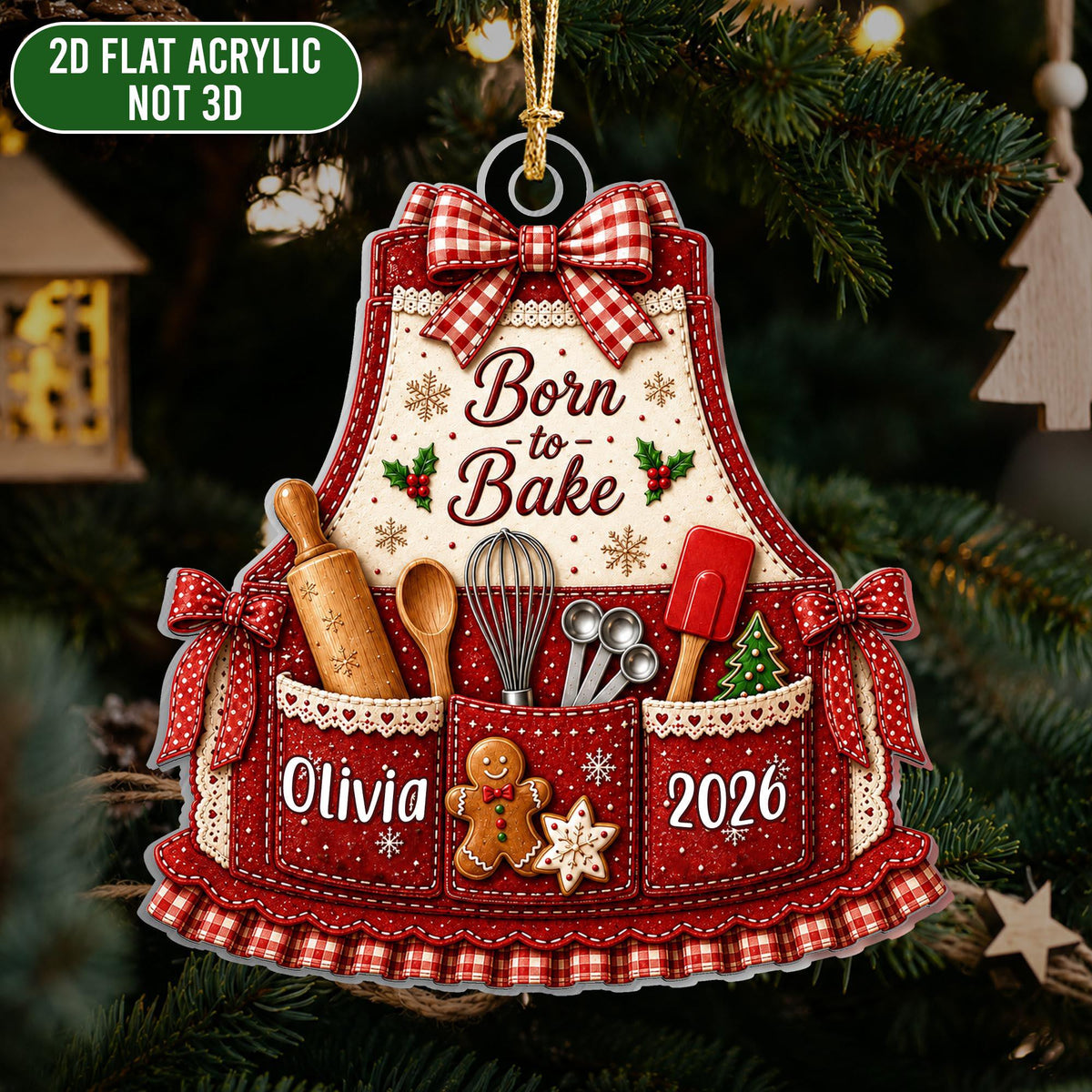 May include: A 2D flat acrylic Christmas ornament shaped like a red and white apron with the text Born to Bake, Olivia, and 2026. The design features a rolling pin, whisk, measuring spoons, spatula, a gingerbread man, and a star cookie.