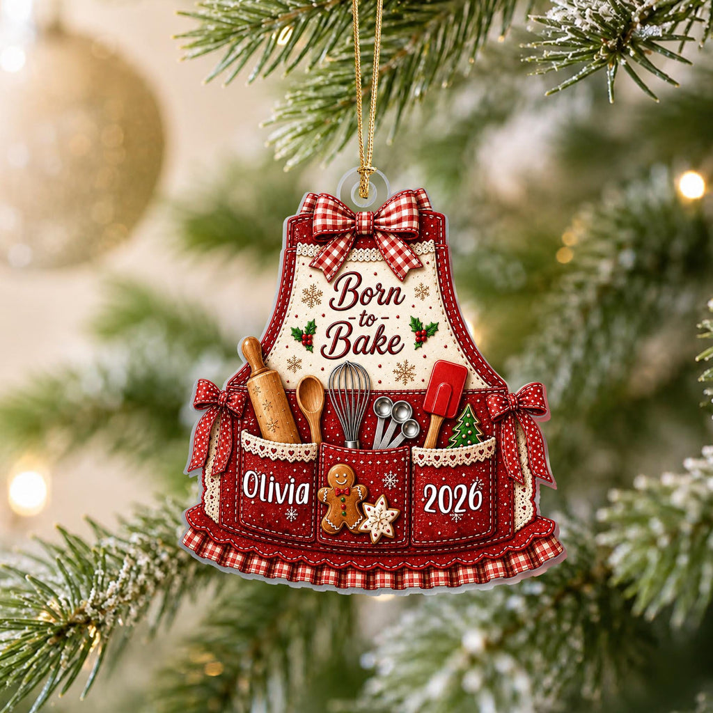 May include: A red and white apron-shaped Christmas ornament featuring a red gingham bow, the text Born to Bake, a gingerbread man, and kitchen utensils. The apron includes the name Olivia and the year 2026 printed on the front pockets.