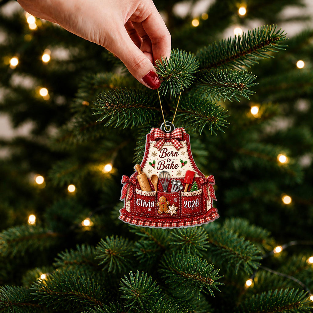 May include: A red and white apron-shaped Christmas ornament featuring a gingham bow, the text "Born to Bake", "Olivia", and "2026". The design includes illustrations of a rolling pin, whisk, measuring spoons, spatula, and a gingerbread man.