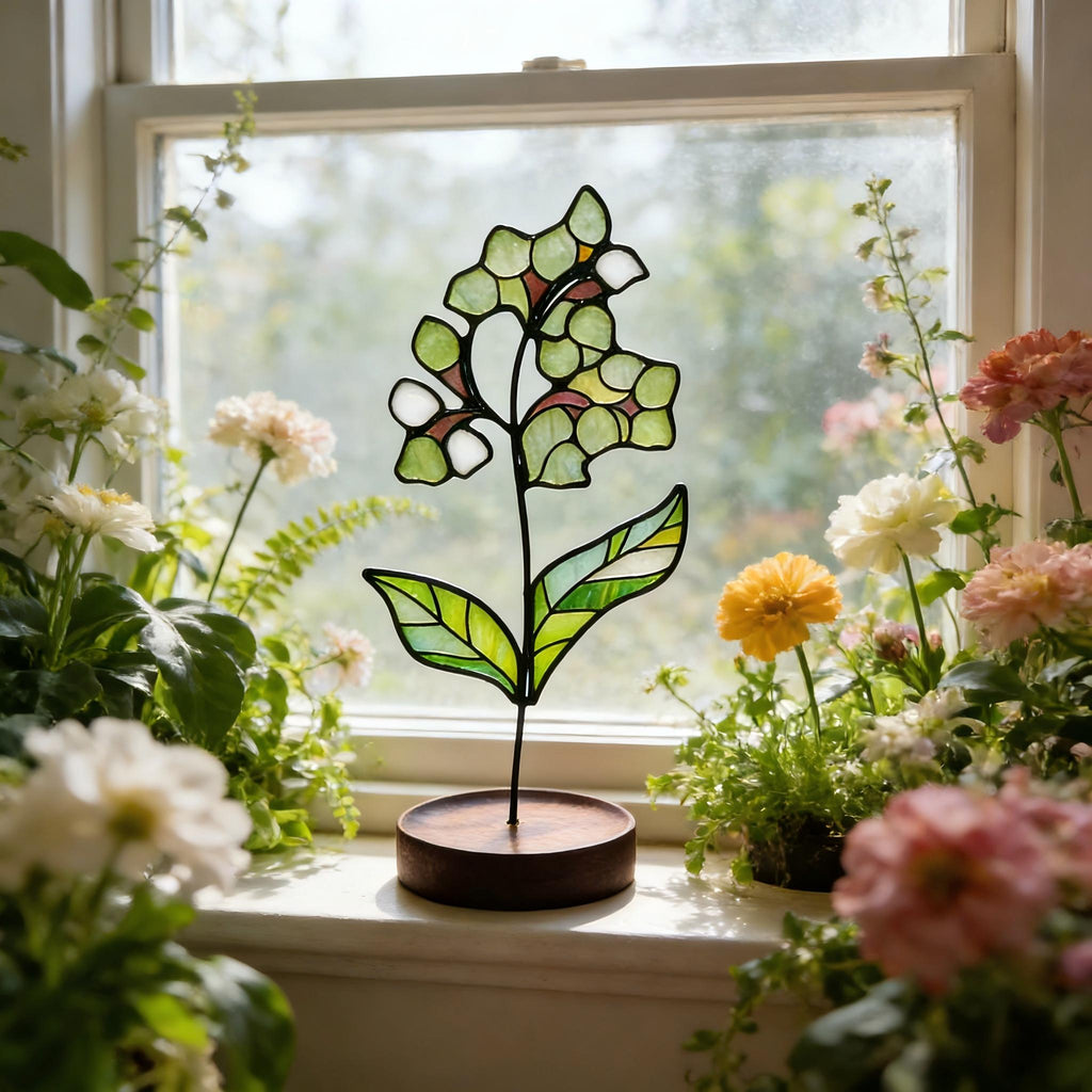 May include: A stained glass flower sculpture featuring translucent green and white glass segments held by black metal cames. The flower is attached to a thin black metal stem that is mounted into a circular, dark brown wooden base.
