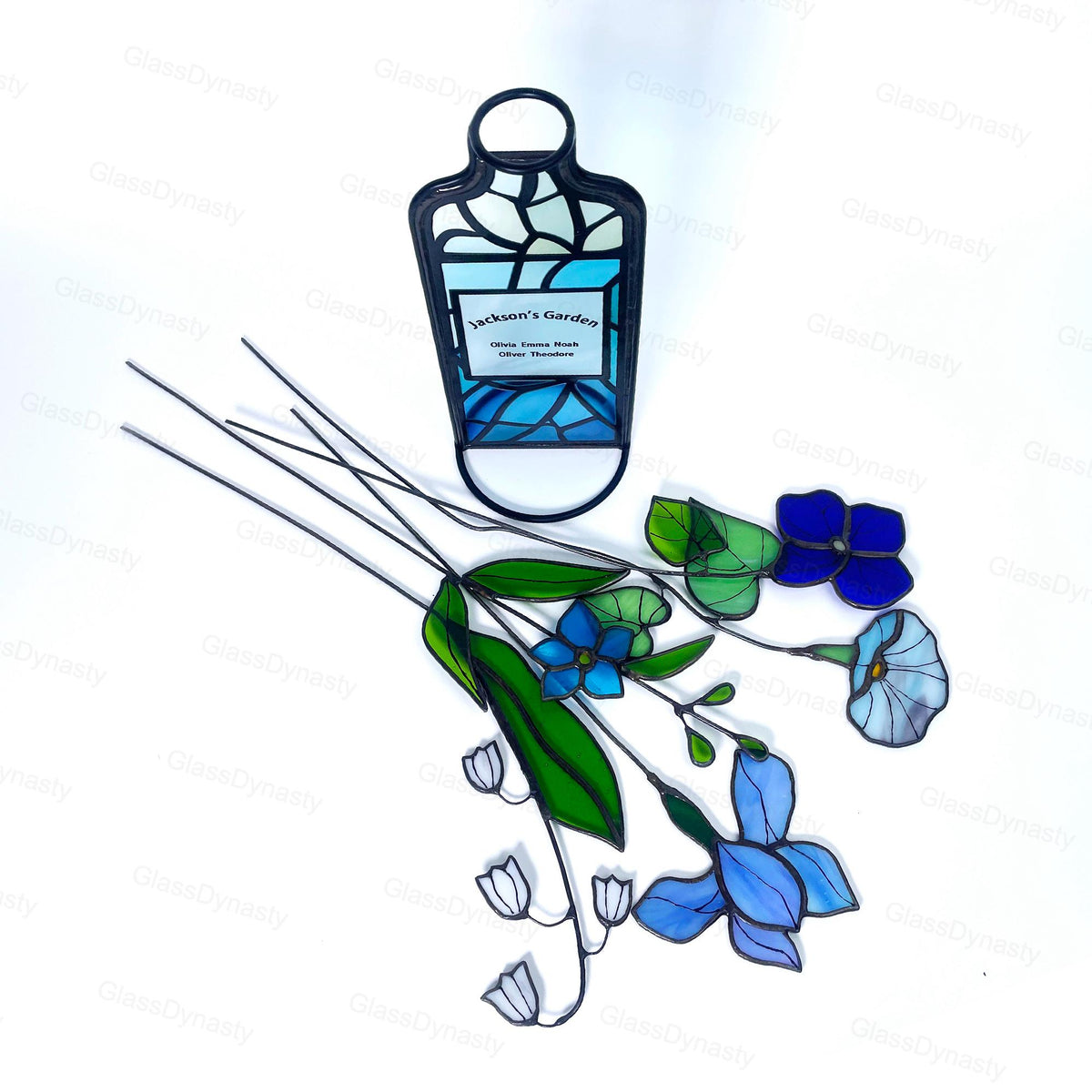 May include: Stained glass art piece featuring a stylized bottle with the text "Jackson's Garden". The bottle is black with blue and white glass. Also, a collection of stained glass flowers in shades of blue, green, and white.