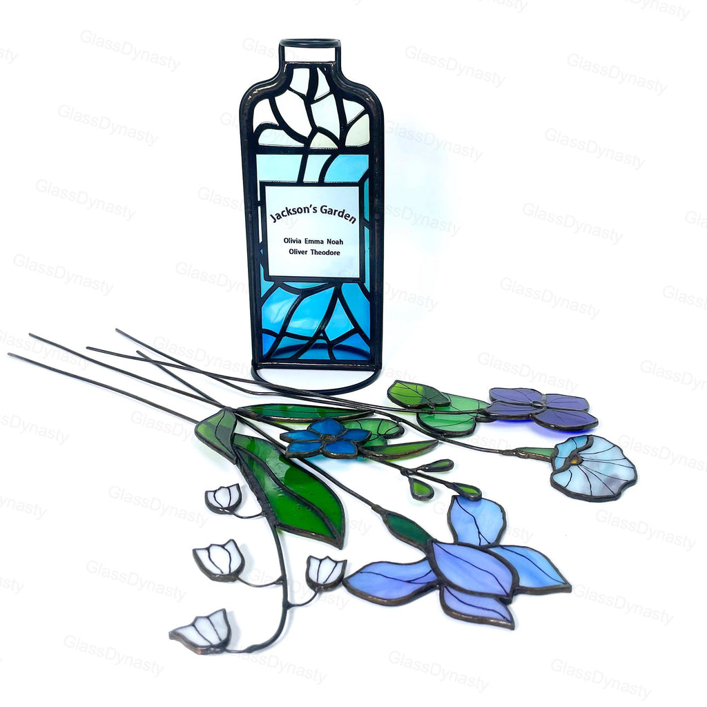 May include: A stained glass garden art piece featuring a bottle-shaped design with the text "Jackson's Garden" and names. The artwork includes blue, green, and purple glass flowers with black stems, creating a decorative garden theme.
