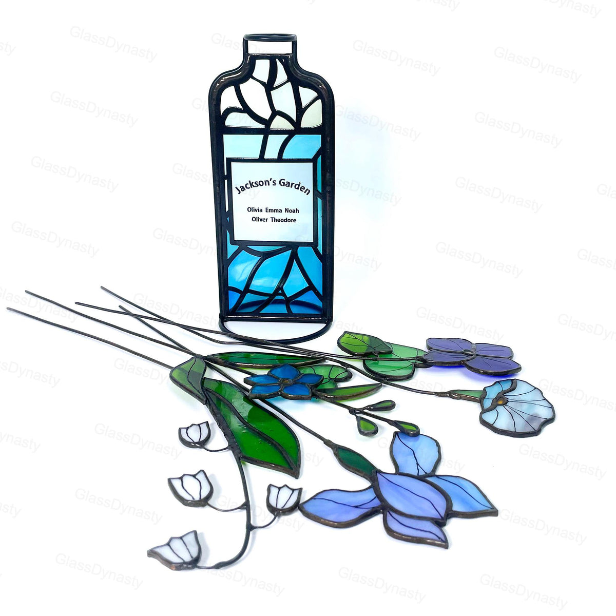 May include: A stained glass garden art piece featuring a bottle-shaped design with the text "Jackson's Garden" and names. The artwork includes blue, green, and purple glass flowers with black stems, creating a decorative garden theme.