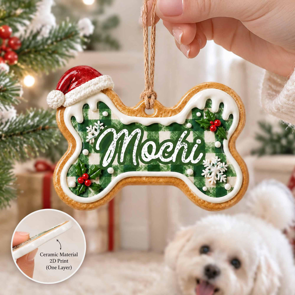May include: A bone-shaped Christmas ornament featuring a green and white buffalo plaid pattern, the name Mochi in white script, a red Santa hat, white icing trim, and holly berry accents. It hangs from a brown twine loop for display.