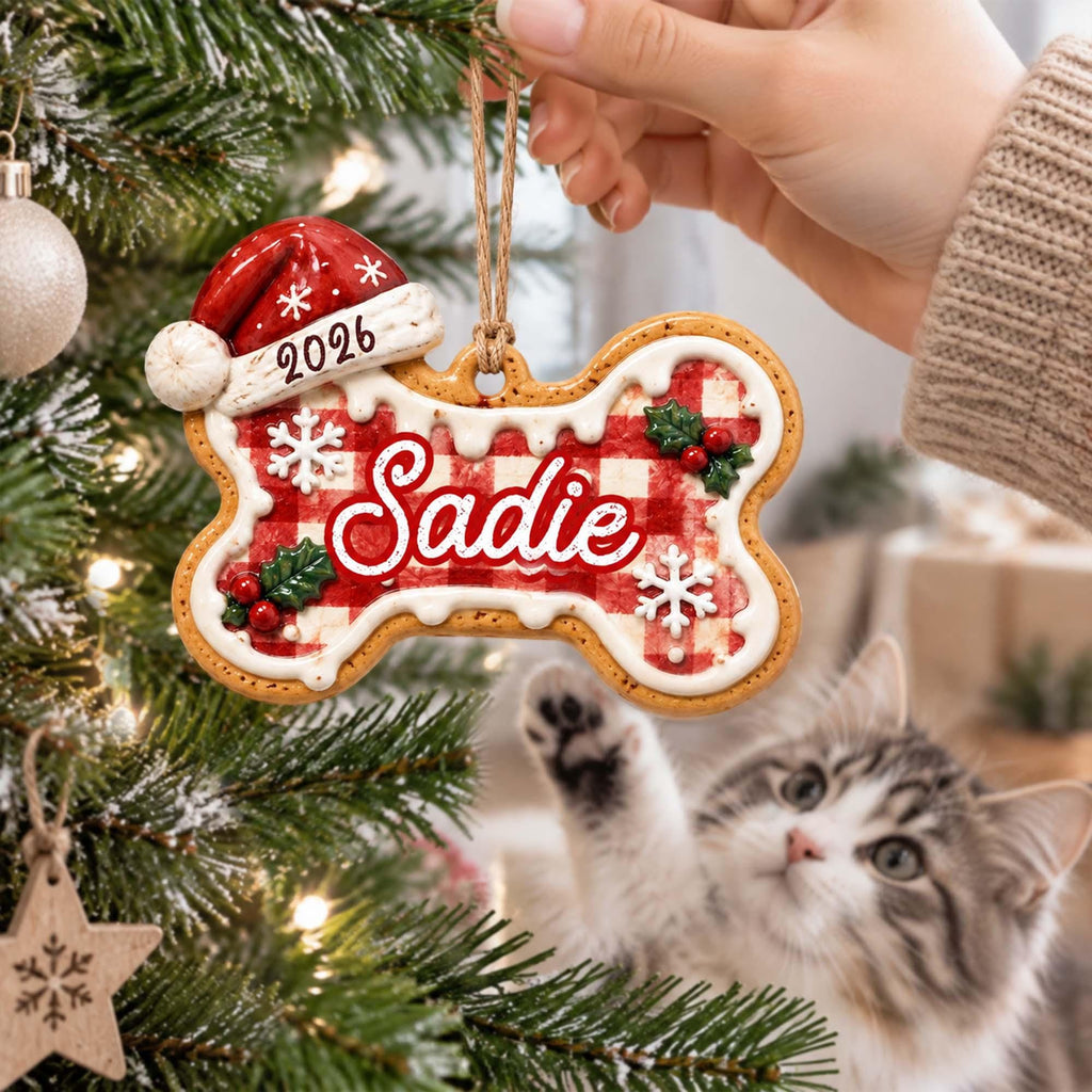 May include: A bone-shaped Christmas ornament featuring a red and white gingham pattern, white icing trim, and holly accents. The name Sadie is written in red script, with the year 2026 on a red Santa hat topper. It hangs from a brown twine loop.