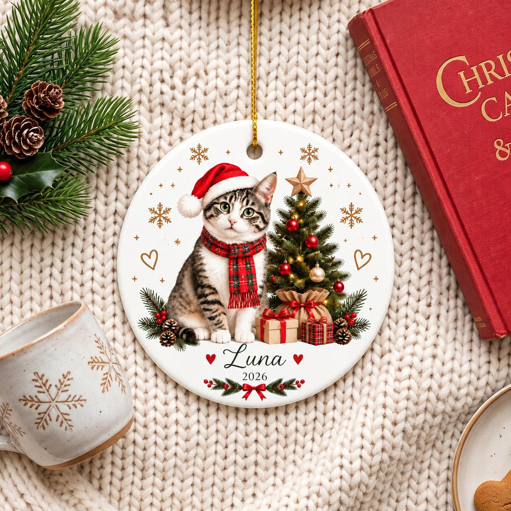 May include: A round white ceramic ornament featuring a tabby cat wearing a red Santa hat and a red plaid scarf. Next to the cat is a decorated Christmas tree with gifts, gold snowflakes, and the text Luna 2026 printed in black with red hearts.