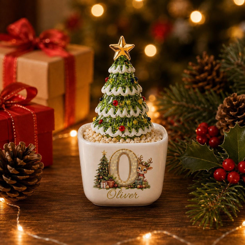 May include: A white ceramic square planter featuring a large letter O, a small reindeer, and a Christmas tree illustration with the name Oliver printed below. A green glass Christmas tree with a gold star sits inside the planter on beige filler.
