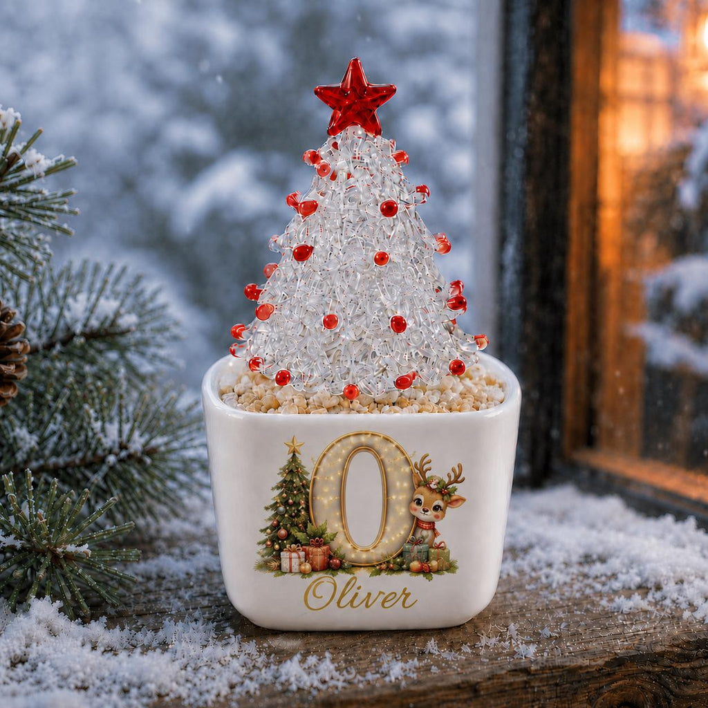May include: A white ceramic square planter featuring a large letter O, a cartoon reindeer, and a Christmas tree illustration with the name Oliver printed below. The planter holds a clear, textured conical Christmas tree topped with a red star.
