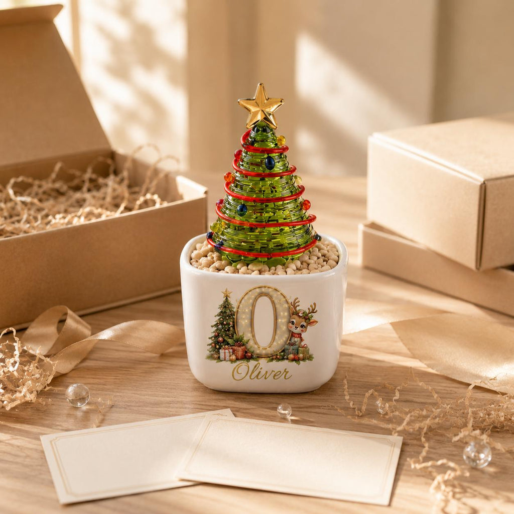 May include: A green glass Christmas tree ornament with a gold star topper, red garland, and colorful beads, set in a white square ceramic planter filled with small stones. The planter features a large letter O, a reindeer, and the name Oliver.