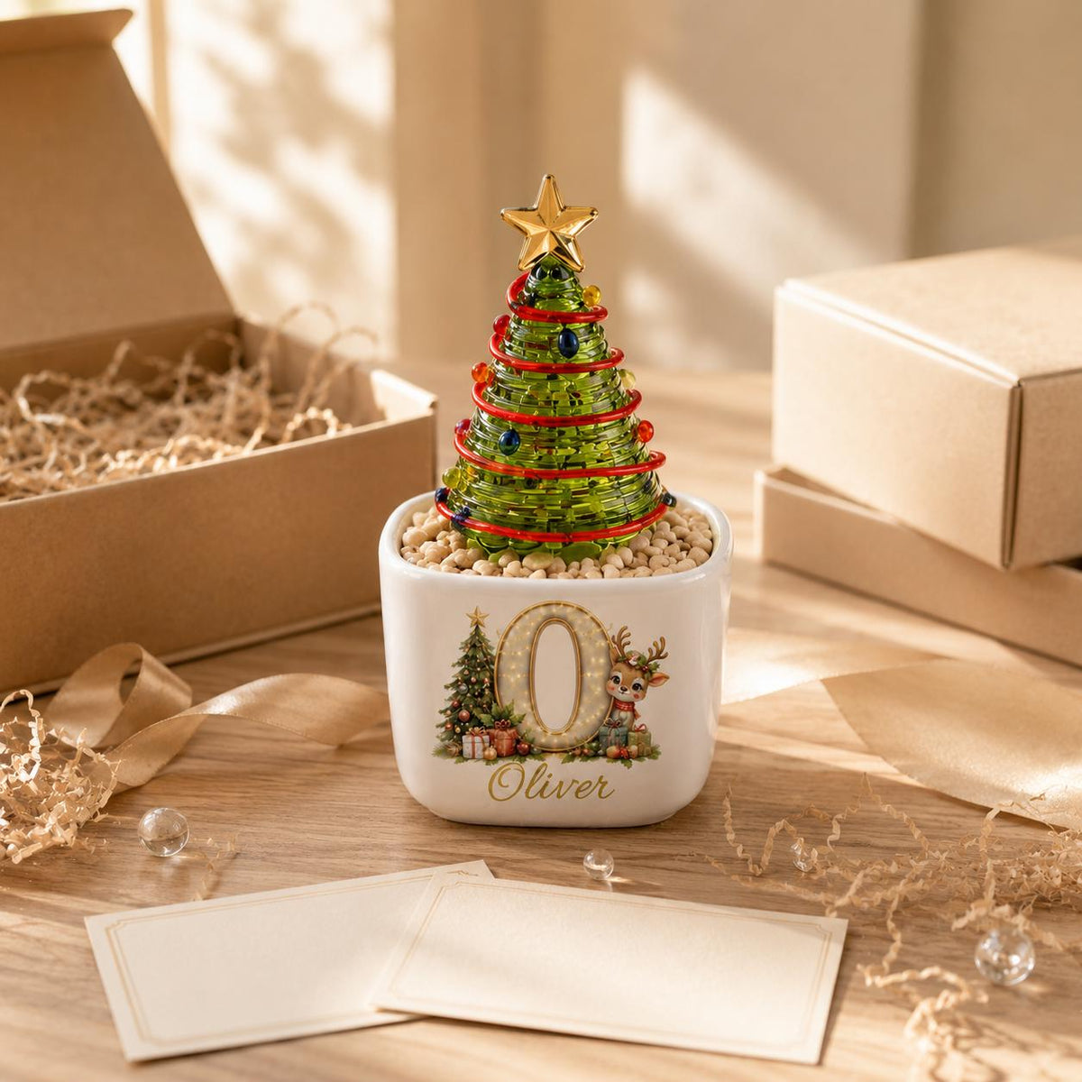 May include: A green glass Christmas tree ornament with a gold star topper, red garland, and colorful beads, set in a white square ceramic planter filled with small stones. The planter features a large letter O, a reindeer, and the name Oliver.