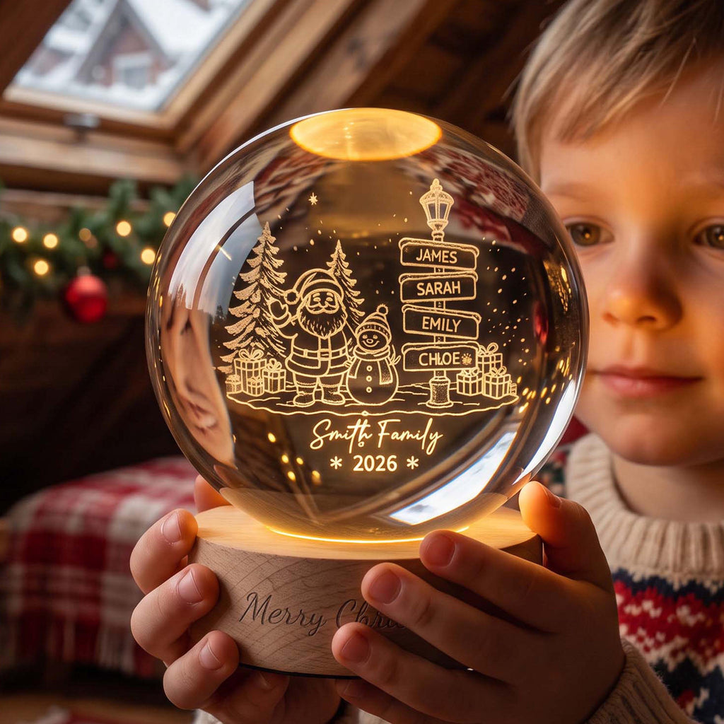 May include: A clear glass sphere night light on a round wooden base. The glass features an etched design of Santa Claus, a snowman, pine trees, gifts, and a street sign with the names James, Sarah, Emily, and Chloe. Text reads Smith Family 2026.
