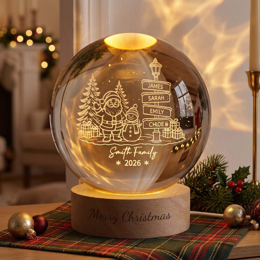 May include: A spherical glass lamp on a light wood base with the text Merry Christmas engraved on the front. The glass features an etched design of Santa, a snowman, pine trees, gifts, and a signpost with the names James, Sarah, Emily, and Chloe.