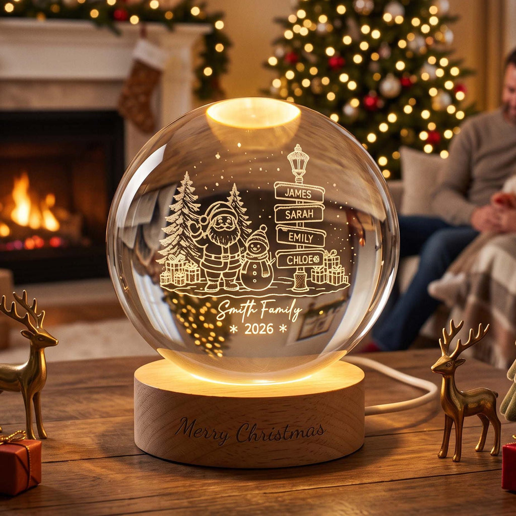 May include: A clear glass sphere lamp on a round wooden base engraved with Merry Christmas. The sphere features an etched design of Santa Claus, a snowman, pine trees, gifts, and a street sign with the names James, Sarah, Emily, and Chloe.