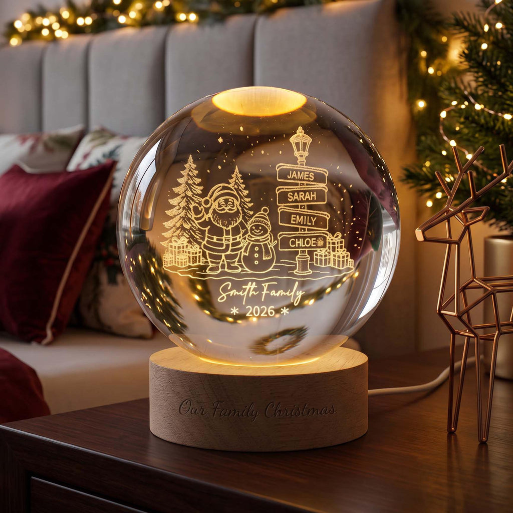 May include: A spherical glass night light on a cylindrical wooden base. The glass features an etched design of Santa Claus, a snowman, pine trees, and a street sign with the names James, Sarah, Emily, and Chloe. Text reads Smith Family 2026.