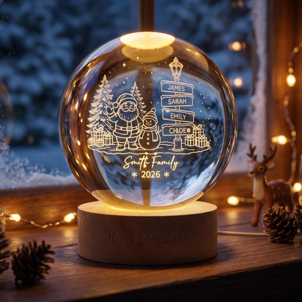 May include: A spherical glass ornament on a round wooden base. The glass features an etched design of Santa Claus, a snowman, pine trees, and a signpost with the names James, Sarah, Emily, and Chloe. It includes the text Smith Family 2026.
