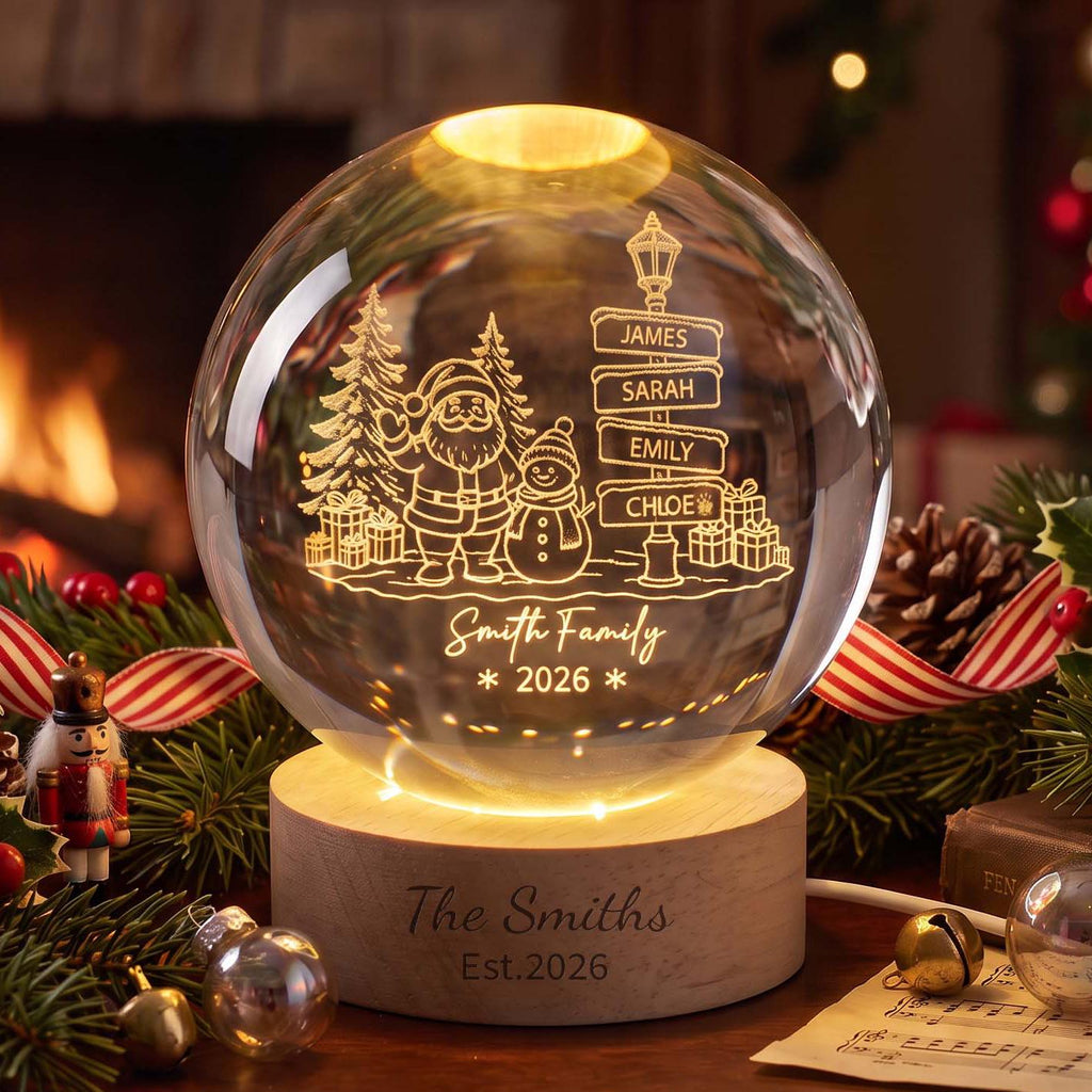May include: A spherical glass ornament with an internal light, featuring an etched design of Santa Claus, a snowman, pine trees, gifts, and a signpost with the names James, Sarah, Emily, and Chloe. The base is wood with The Smiths Est. 2026 engraved.