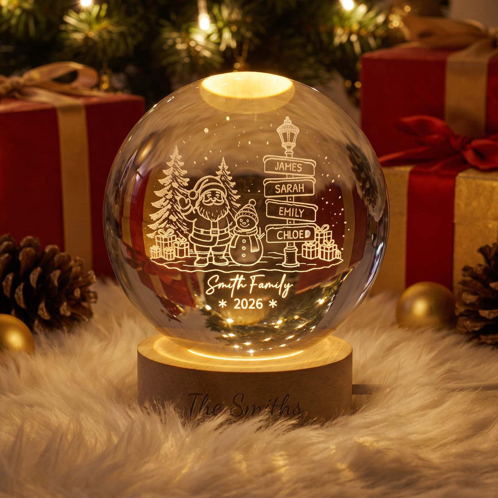 May include: A spherical glass night light on a wooden base featuring an etched design of Santa Claus, a snowman, pine trees, and a signpost with the names James, Sarah, Emily, and Chloe. The text Smith Family and 2026 are engraved below.