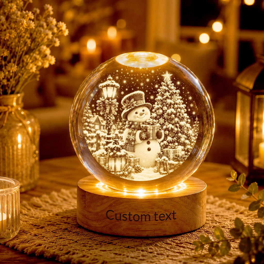 May include: A clear glass globe ornament featuring an internal 3D laser-etched scene of a snowman wearing a hat and scarf, standing next to a decorated Christmas tree and a street lamp. The globe rests on a round wooden base with the text Custom text.