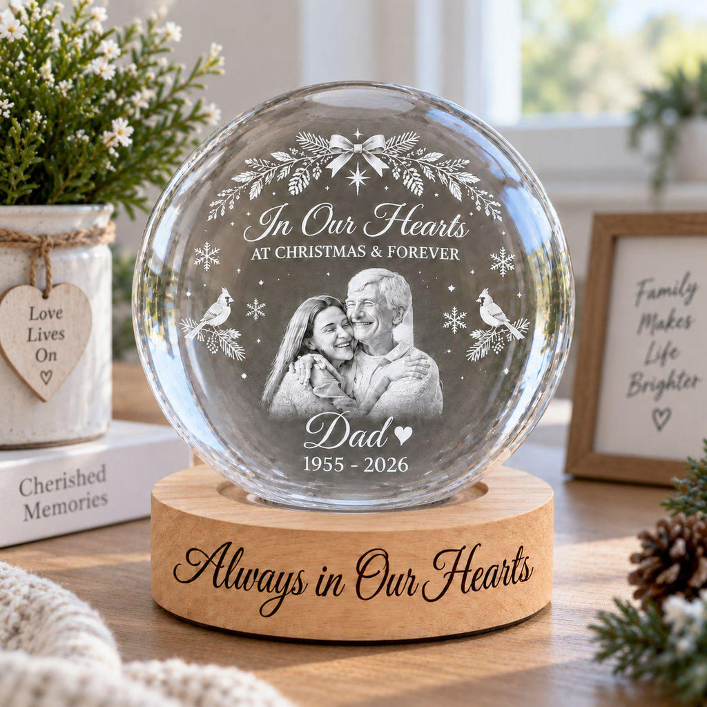May include: A clear glass sphere ornament on a round wooden base. The glass features an etched image of a couple, a wreath, snowflakes, and two birds. Text reads: In Our Hearts AT CHRISTMAS & FOREVER, Dad, 1955 - 2026. The base reads: Always in Our Hearts.