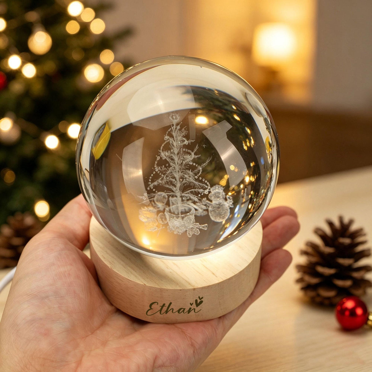 May include: A clear glass sphere featuring an internal laser-etched design of a Christmas tree and a snowman. The sphere rests on a round, light-colored wooden base that is illuminated from below and engraved with the name Ethan and a small heart.