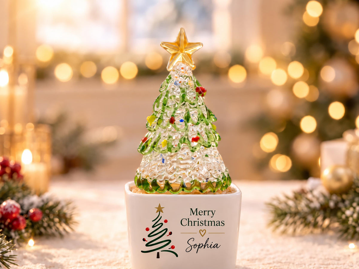 May include: A clear glass Christmas tree figurine with green branches and multi-colored ornaments, topped with a gold star. The tree sits in a white square ceramic pot featuring a printed green tree, the text Merry Christmas, and the name Sophia.
