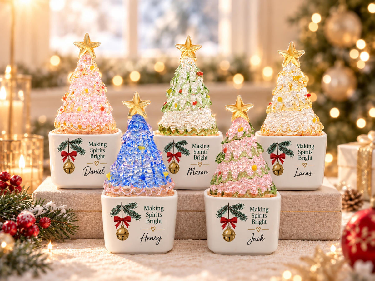 May include: Five decorative tabletop Christmas trees with textured, translucent, colorful glass-like foliage topped with gold stars. Each tree sits in a white square ceramic base featuring a pine branch, a red bow, a gold bell, and the text Making Spirits Bright with a personalized name.