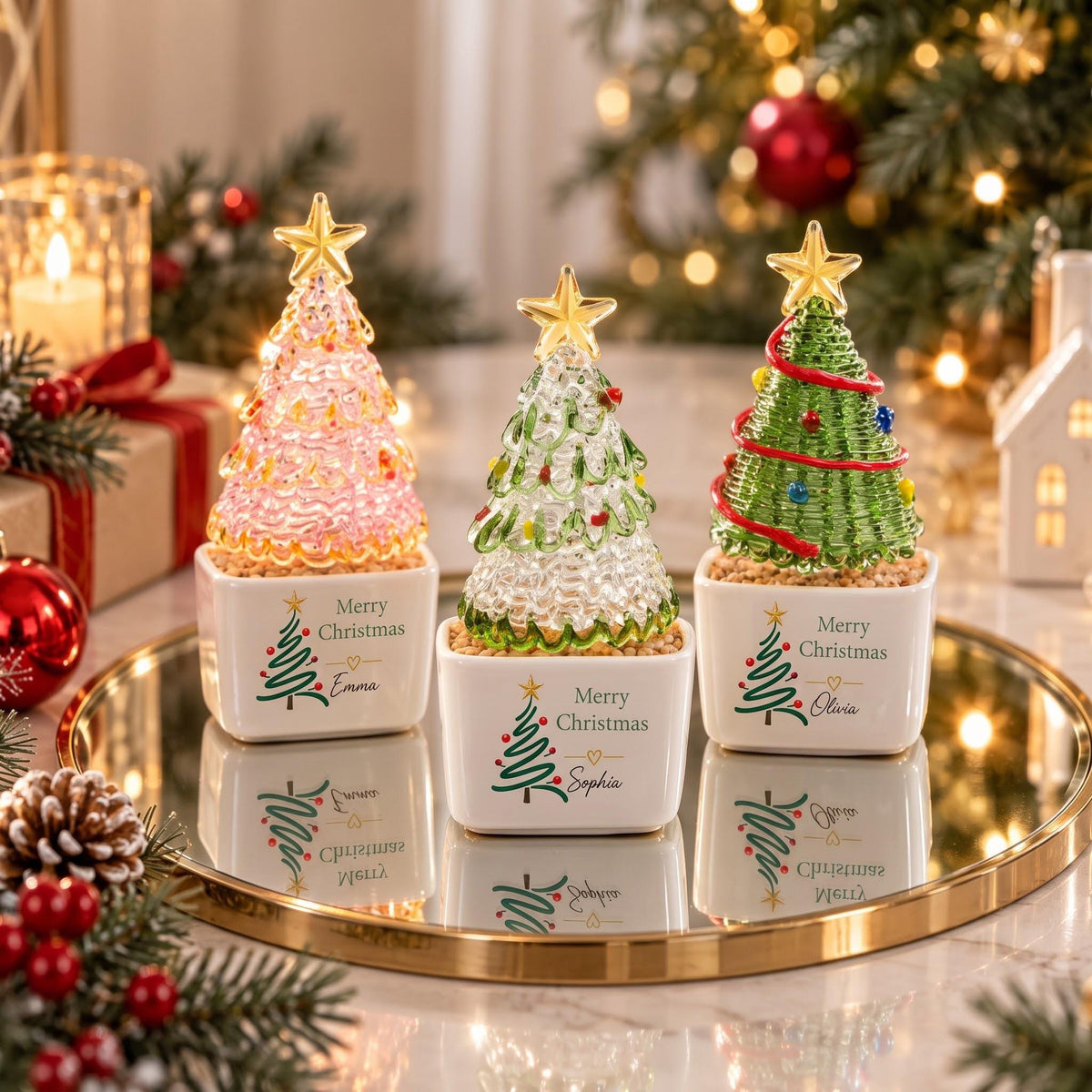 May include: Three glass Christmas tree ornaments in white square ceramic pots. Each tree is topped with a gold star and features colorful accents. The pots are printed with a green tree graphic, the text Merry Christmas, and names Emma, Sophia, or Olivia.