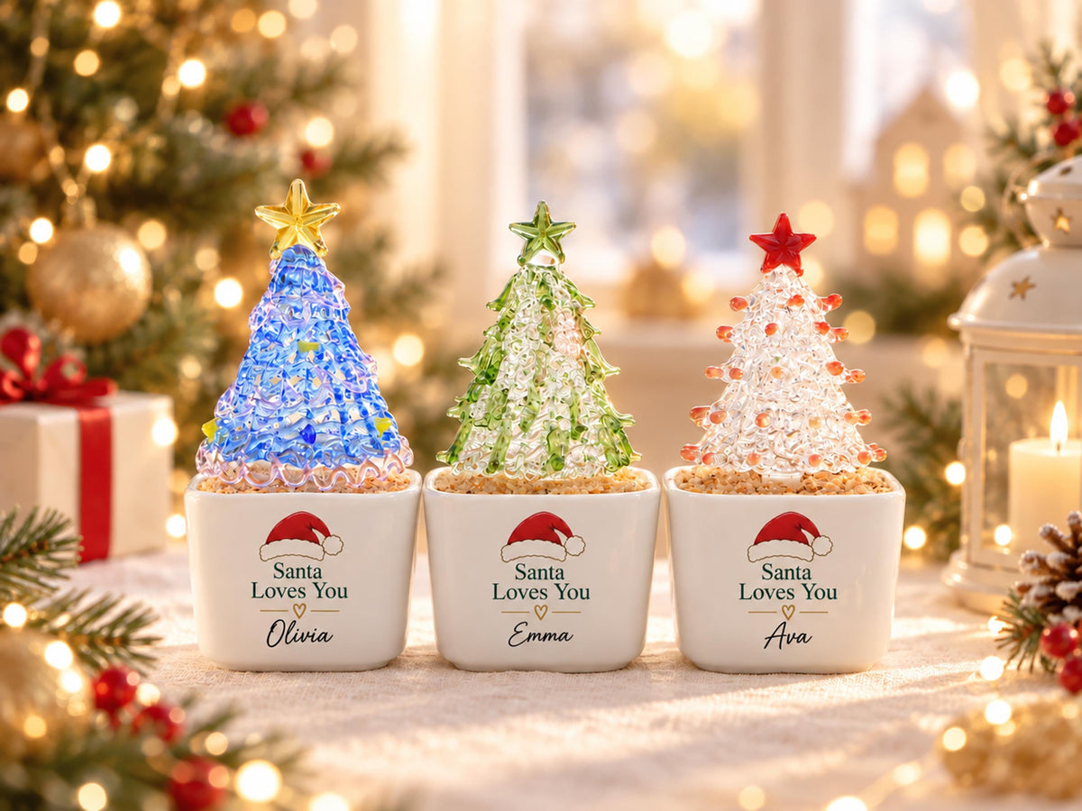 May include: Three small decorative glass Christmas trees in white square ceramic pots. Each pot features a red Santa hat graphic, the text "Santa Loves You" with a heart, and a name: Olivia, Emma, or Ava. The trees are blue, green, and clear.