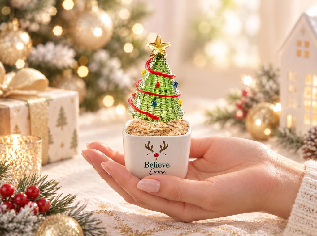 May include: A small green wire Christmas tree ornament with a gold star topper, red garland, and colorful beads, set in a white square ceramic pot featuring a reindeer face graphic, the word Believe, and the name Emma printed in black.