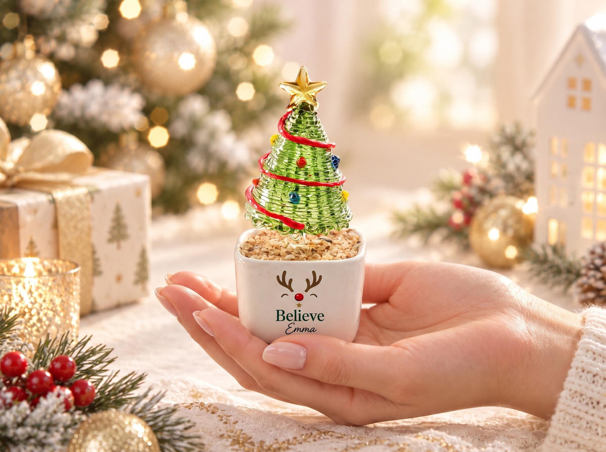 May include: A small green wire Christmas tree ornament with a gold star topper, red garland, and colorful beads, set in a white square ceramic pot featuring a reindeer face graphic, the word Believe, and the name Emma printed in black.