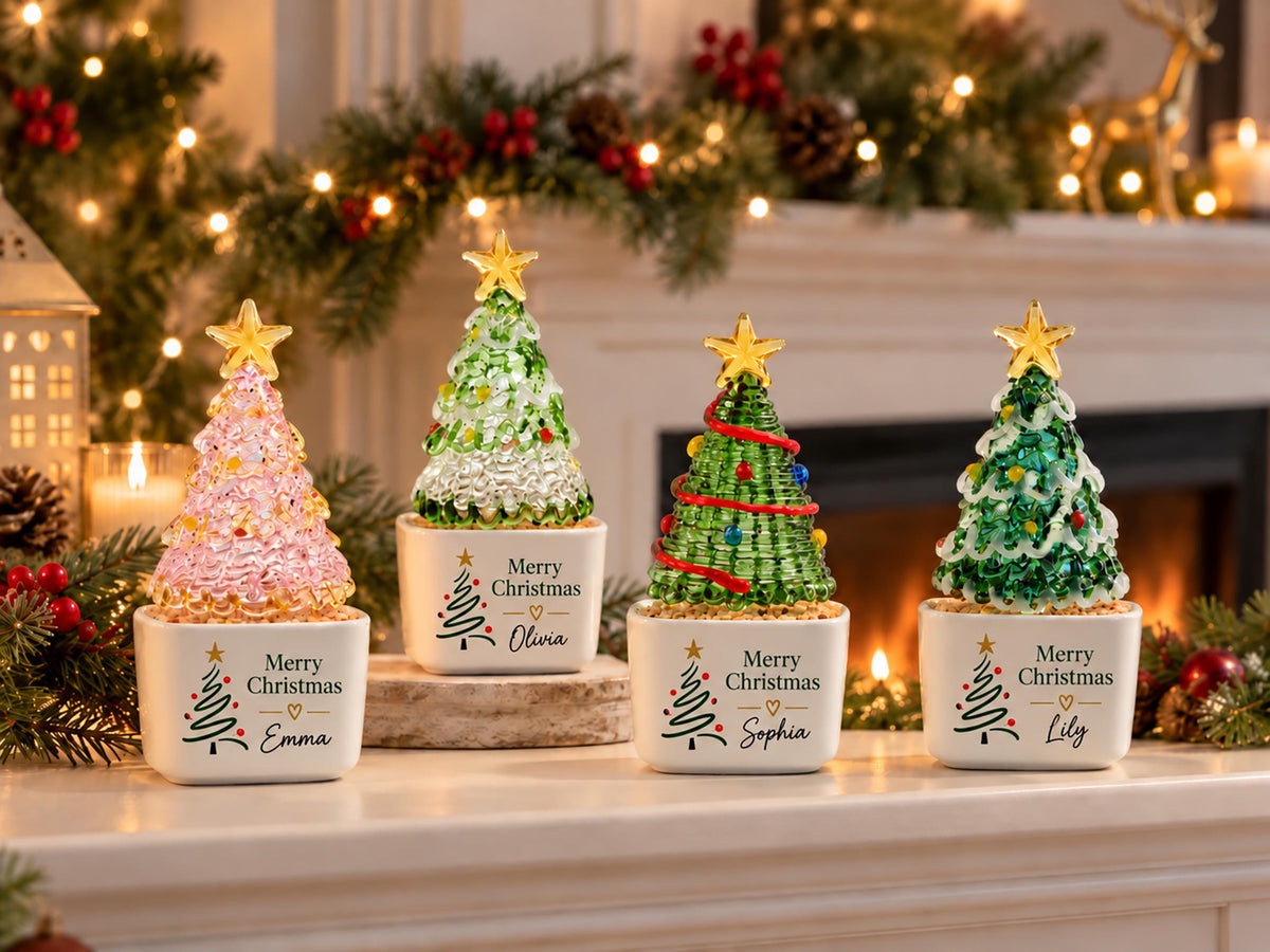 May include: Four decorative glass Christmas tree figurines in white ceramic pots. Each pot features the text Merry Christmas, a small heart, and a name: Emma, Olivia, Sophia, or Lily. The trees are topped with gold stars and have colorful accents.