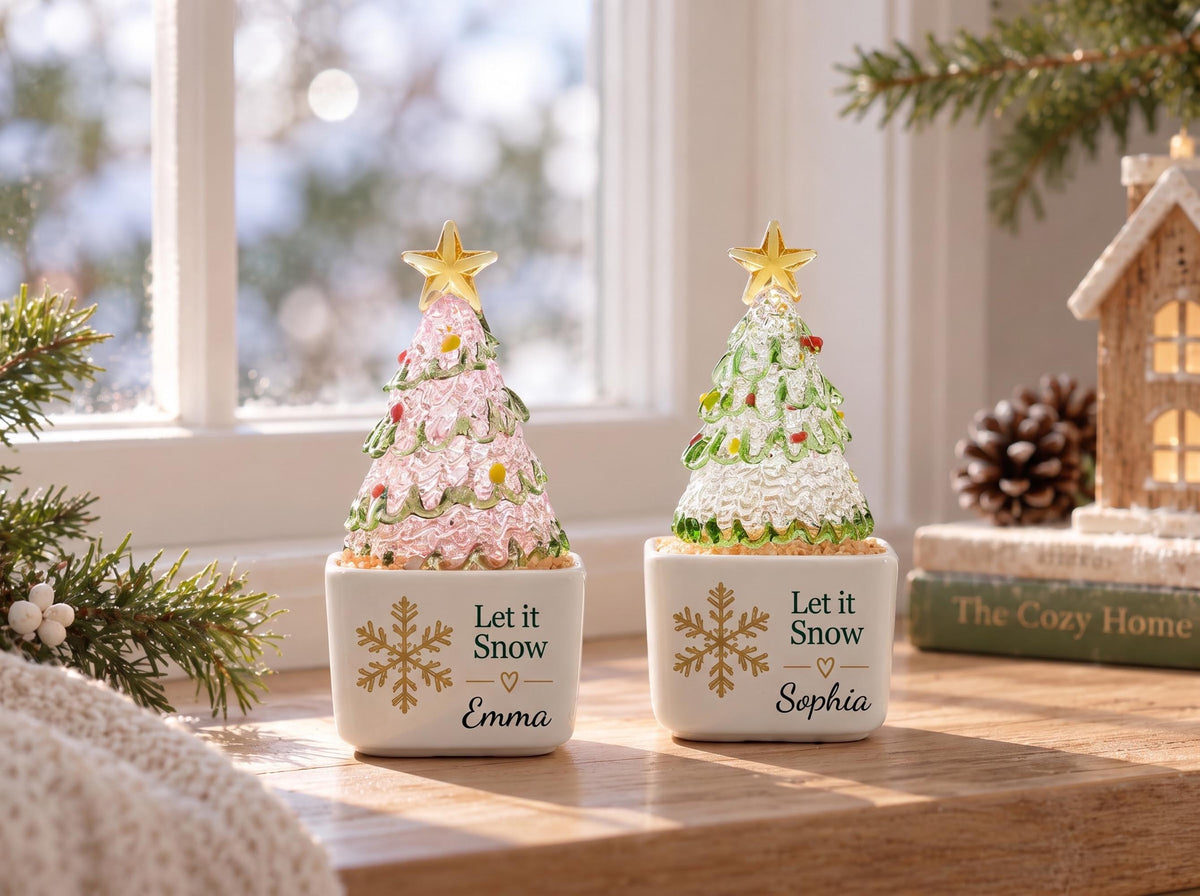 May include: Two decorative glass Christmas trees in white ceramic square pots. One tree is pink, the other green, both topped with a gold star. Each pot features a gold snowflake, the text Let it Snow, a small heart, and a name, Emma or Sophia.