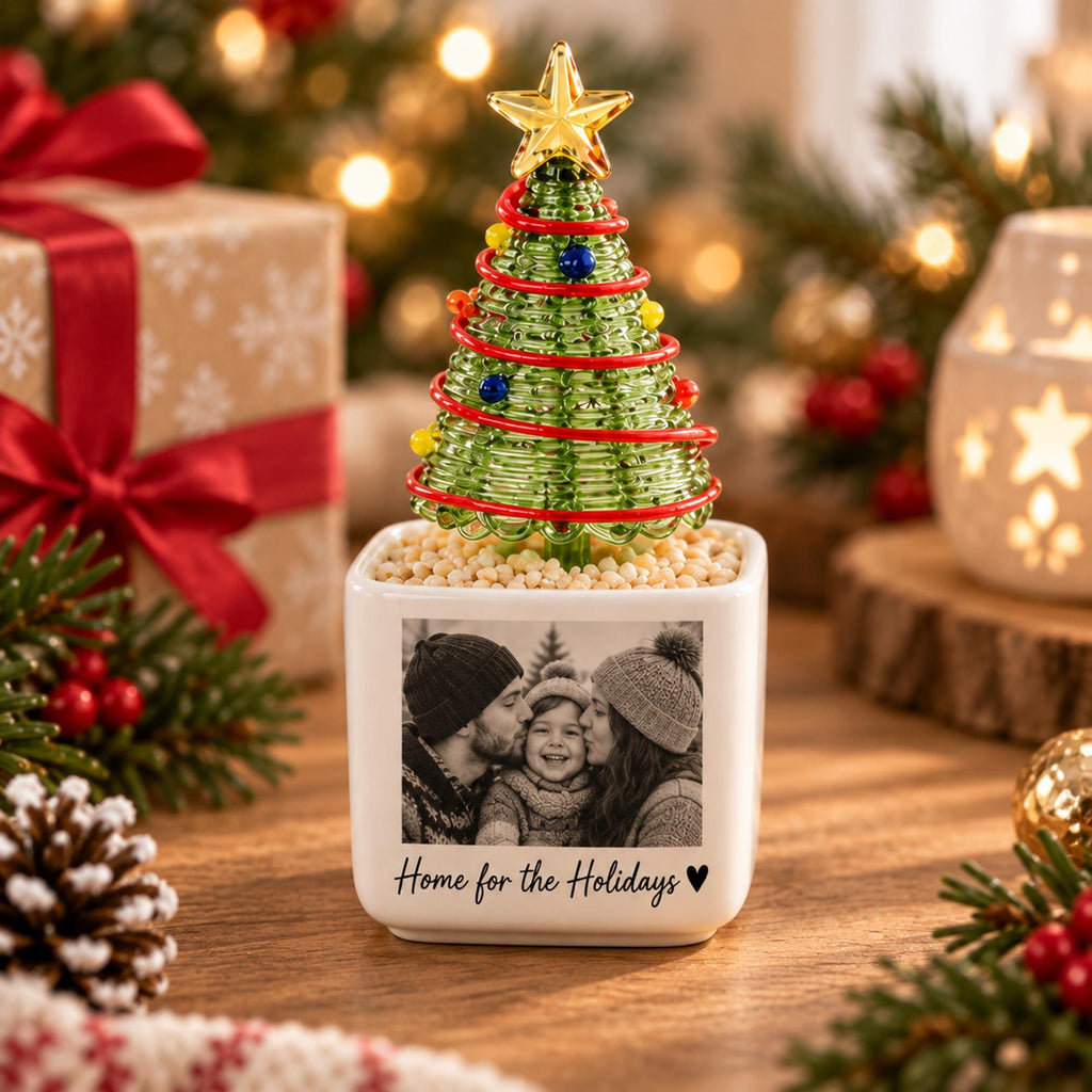 May include: A white square ceramic planter featuring a black and white photograph of a family and the text Home for the Holidays with a heart symbol. A green glass Christmas tree with a gold star topper sits in the planter on beige filler.