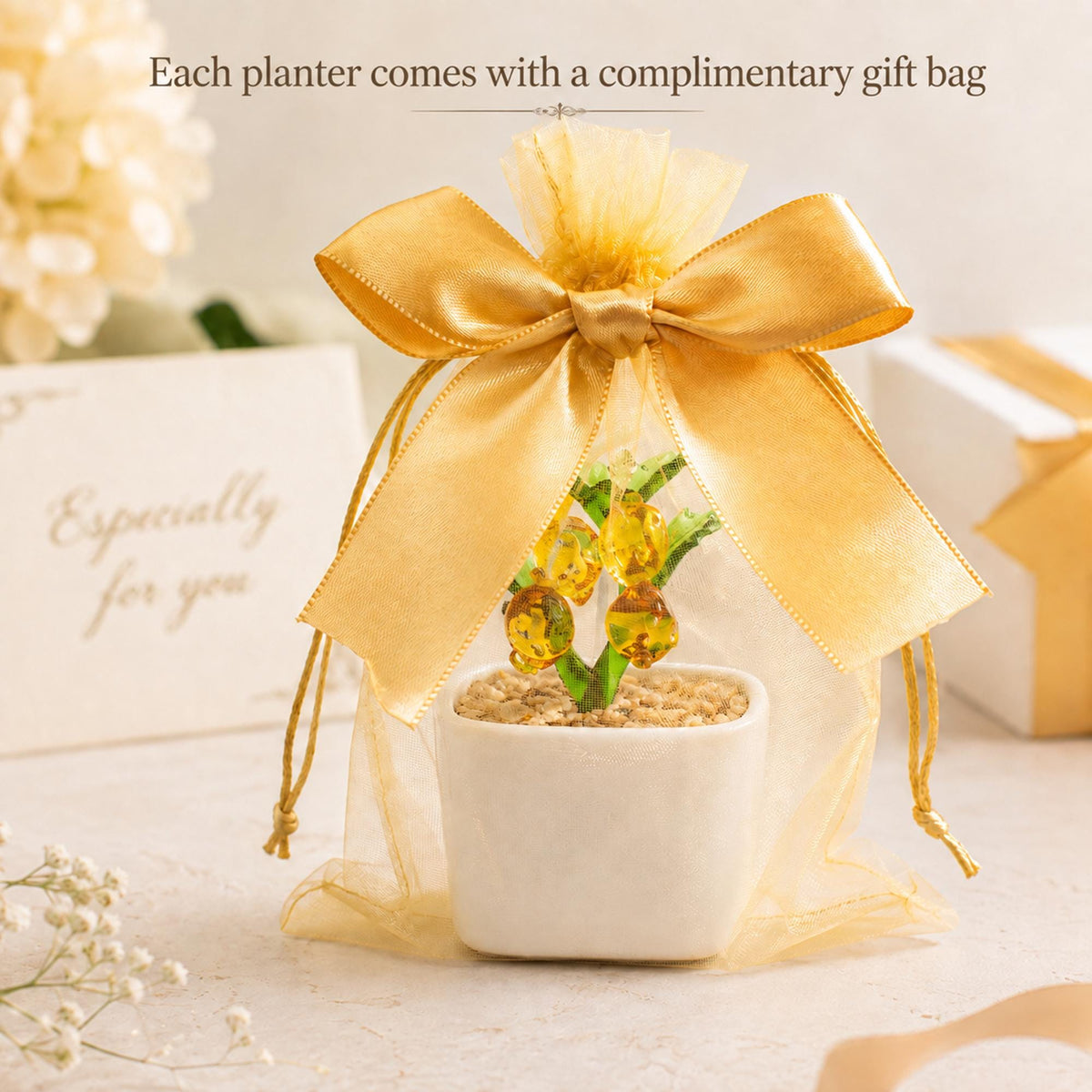 May include: A small white square planter containing a green plant with amber-colored glass fruit, encased in a sheer gold organza drawstring gift bag topped with a large satin gold ribbon bow. The text "Each planter comes with a complimentary gift bag" is visible.