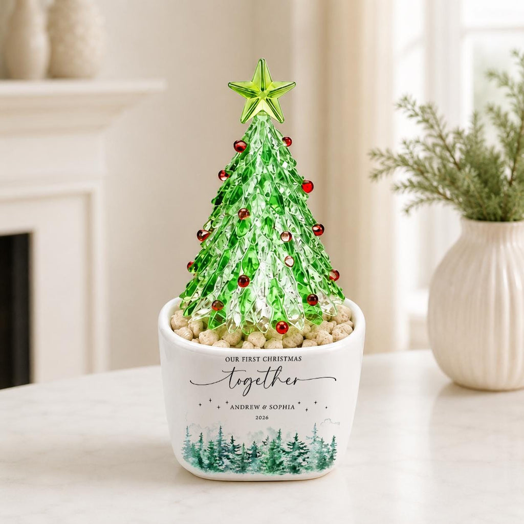 May include: A glass Christmas tree ornament with green foliage and red accents, topped with a green star. It sits in a white ceramic pot featuring the text "OUR FIRST CHRISTMAS together ANDREW & SOPHIA 2026" and a green forest graphic.