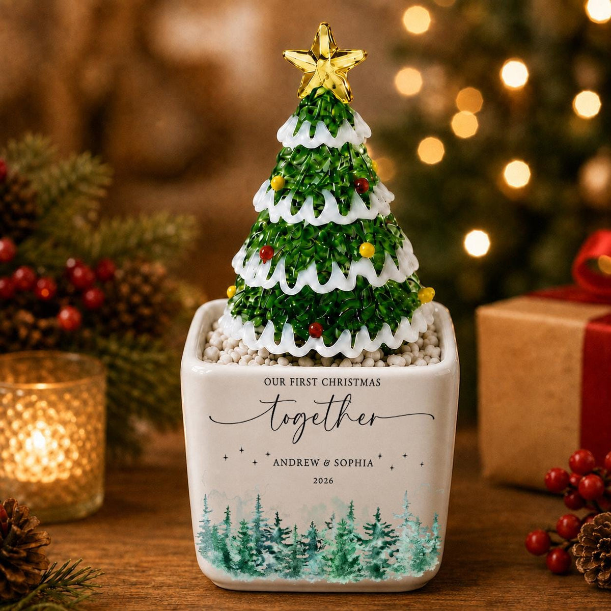 May include: A decorative glass Christmas tree with green branches, white snow accents, and a gold star topper, set in a white square ceramic planter. The planter features the text "OUR FIRST CHRISTMAS together" and "ANDREW & SOPHIA 2026" above a green forest graphic.