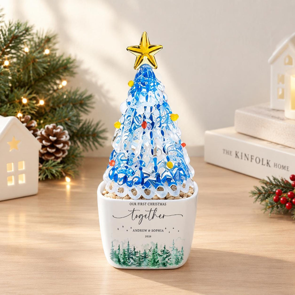 May include: A decorative glass Christmas tree ornament with blue and white swirling patterns, topped with a gold star and small colorful beads. It sits in a white square ceramic planter featuring the text "OUR FIRST CHRISTMAS together ANDREW & SOPHIA 2024" and a green forest graphic.