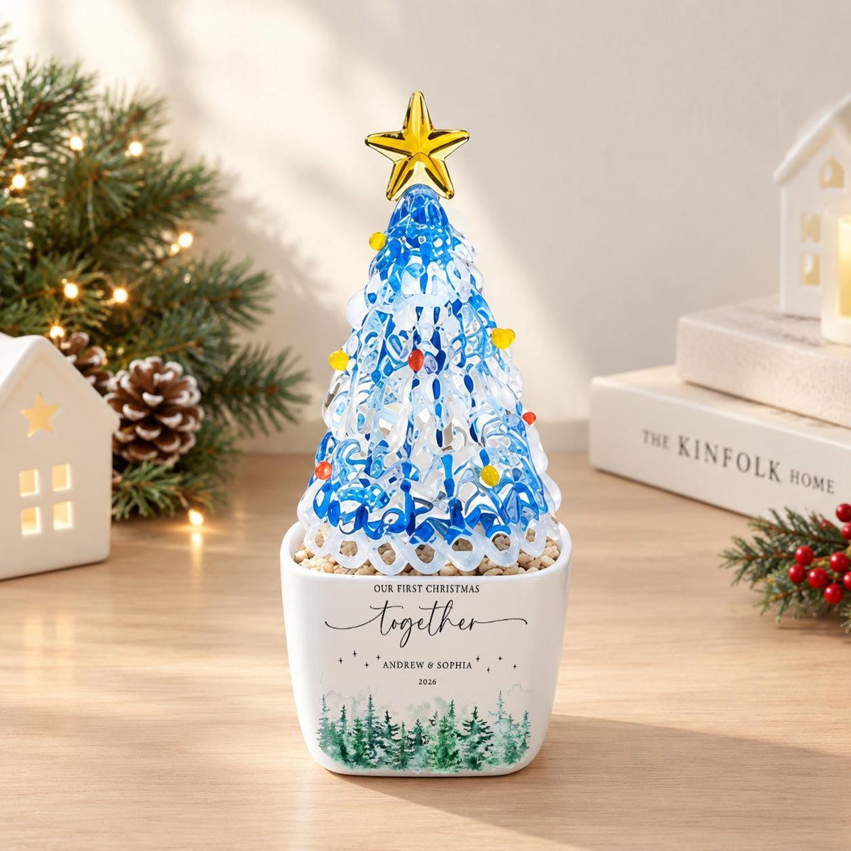 May include: A decorative glass Christmas tree ornament with blue and white swirling patterns, topped with a gold star and small colorful beads. It sits in a white square ceramic planter featuring the text "OUR FIRST CHRISTMAS together ANDREW & SOPHIA 2024" and a green forest graphic.