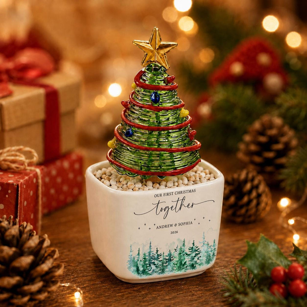 May include: A miniature Christmas tree made of green wire wrapped in red wire with small colorful beads, topped with a gold star. It sits in a white square ceramic pot featuring the text "OUR FIRST CHRISTMAS together" and "ANDREW & SOPHIA 2026" above a painted green forest scene.