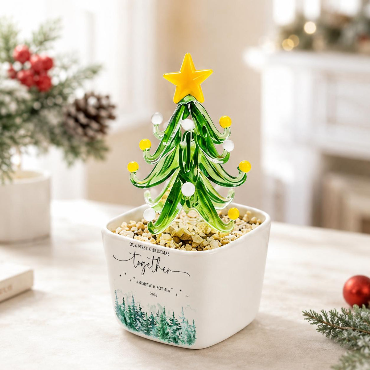May include: A glass Christmas tree ornament with green branches, yellow and white spherical accents, and a yellow star topper. It is set in a white square ceramic planter filled with small stones, featuring the text "OUR FIRST CHRISTMAS together ANDREW & SOPHIA 2036" and a green forest graphic.